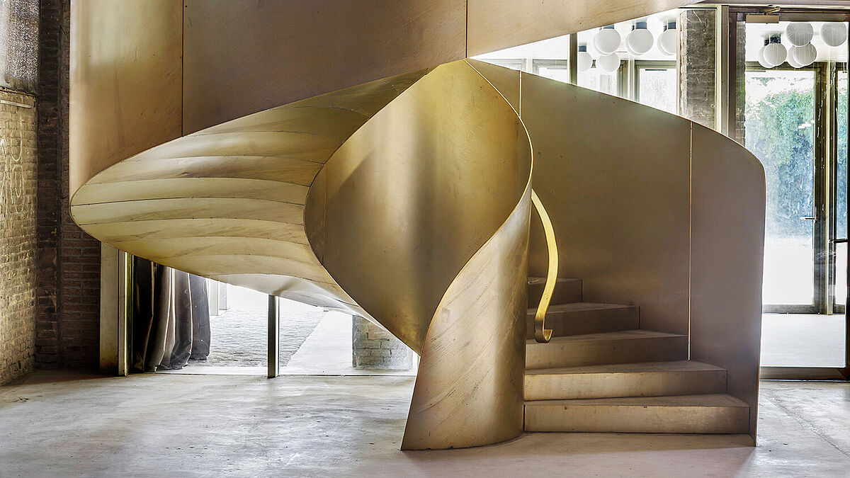 In der offenen Ausstellungshalle führt eine golden glänzende Wendeltreppe mit massiven Blechwangen zum Obergeschoss. 