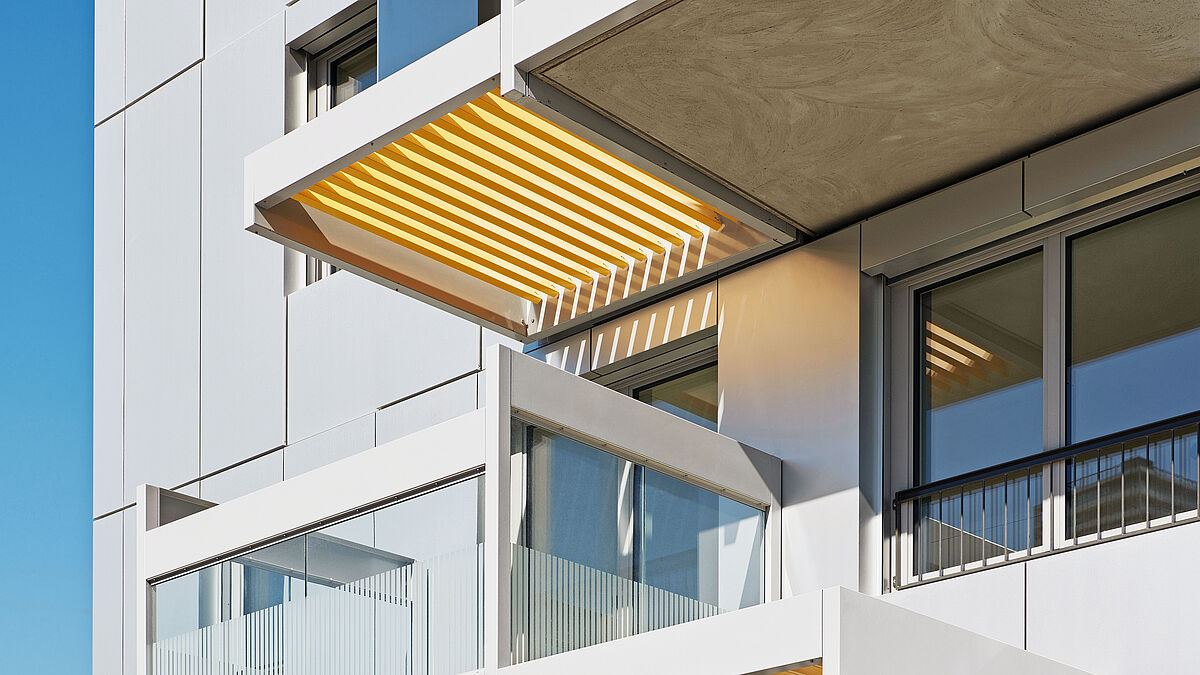 Links die Balkonverkleidungen mit verglastem Geländeraufbau. Rechts die Schattendächer aus bunten Metalllamellen.