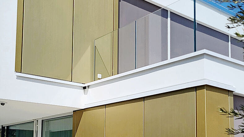 Eine gelungene optische Kombination zwischen der beigen verputzten Fassade, den goldenen Aluminiumblechen und dem kristallfarbenen Glas. 