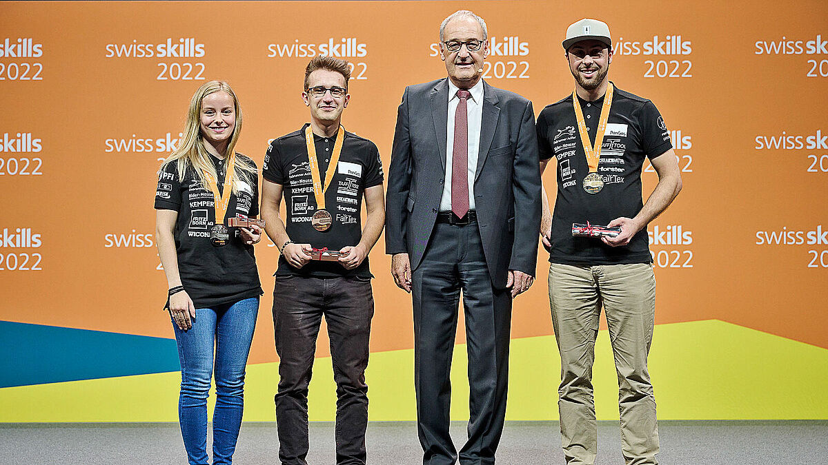 Strahlende Gesichter bei den drei erstplatzierten Metallbaukonstrukteur*innen. Von links: Svenja Knecht (2.), David Fuchs (3.) und  Adrian Gilgen (1.)., zusammen mit   Gratulant Bundesrat Guy Parmelin.