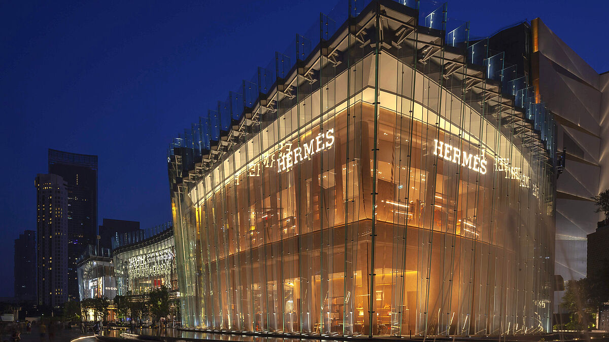 Eine im gleichen Winkel geneigte, durchgehende Innenfassade aus bronzefarbig lackiertem Metallgewebe verleiht dem Hermès-Flagship-Store in Bangkok besonderen Glanz.