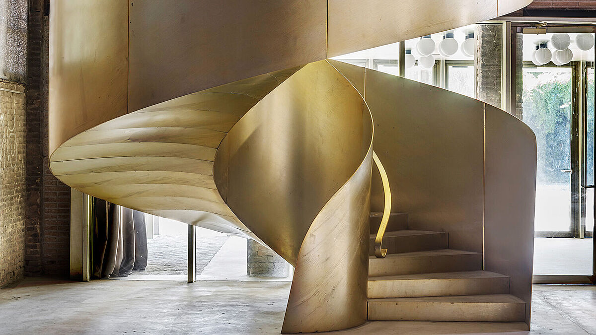 Dans le hall d’exposition ouvert, un escalier en colimaçon doré brillant doté de barres de tôle massives mène à l’étage supérieur. 